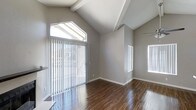 This single-floor space makes a strong first impression with its dramatic vaulted ceilings accented by exposed decorative beams, creating a sense of openness and height that defines the overall atmosphere. The main living area is bathed in natural light, courtesy of a large sliding glass door fitted with vertical blinds and flanked by transom windows that draw the eye upward toward the soaring roofline. Additional windows along the adjacent wall further enhance the brightness and airy feel of the space. Smooth, neutral gray walls provide a clean, contemporary backdrop that complements the warm tones of the rich, dark hardwood flooring, which runs throughout and adds a sense of elegance and warmth. White baseboards and trim offer a crisp contrast, giving the interior a polished, well-finished look. The open layout promotes a flexible use of the space, while the high ceiling contributes to a feeling of expansiveness well beyond what the footprint alone might suggest. The combination of modern finishes, natural light, and architectural detailing makes this a welcoming and visually compelling space suited to a variety of uses.