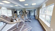 This fitness center offers a comprehensive workout environment designed to support a range of exercise needs. The main cardio area is lined with a row of treadmills and elliptical machines, all positioned along a full-length mirrored wall that enhances the sense of space and allows users to monitor their form during workouts. The flooring throughout is a durable dark carpet, well-suited for a high-traffic exercise environment. Natural light filters in through multiple large windows along the side wall, creating a bright and welcoming atmosphere. Crown molding along the ceiling and neutral sage-toned walls give the space a refined, polished appearance. Recessed flush-mount ceiling lighting ensures consistent illumination throughout. A second area is visible through a connecting opening, housing additional strength and resistance training equipment, expanding the facility's workout options. The space is thoughtfully laid out to allow for comfortable movement between machines and equipment. Overall, this fitness center combines practical functionality with a clean, well-maintained aesthetic, making it an inviting space for both casual users and dedicated fitness enthusiasts.