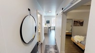 This single-floor unit showcases a contemporary design with a neutral color palette, dark hardwood-style flooring, and recessed lighting throughout. The entry hallway sets a welcoming tone, featuring a round black-framed mirror and a sliding barn door that leads to the bedroom, adding a stylish industrial accent.

The kitchen is efficiently designed with light wood cabinetry, white countertops, and a subtle tile backsplash. Stainless steel appliances—including a refrigerator, range, and microwave—line one wall, while a peninsula with pendant lighting provides additional prep space and casual dining. The open layout connects seamlessly to the living area.

The living room is bright and comfortable, benefiting from large windows that frame urban cityscape views, allowing ample natural light. The space comfortably accommodates a sectional sofa, lounge chair, and entertainment setup.

The bedroom is a calm retreat featuring a ceiling fan, wall-mounted reading sconces, a built-in closet, and a full-length mirror. The clean white walls create a restful atmosphere.

The bathroom maintains the unit's modern aesthetic with light tile flooring, a floating vanity with white countertop, and a tub-shower combination, keeping the overall feel bright and functional.