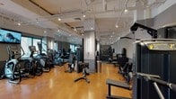 The fitness center at 1 Alexander St North in Yonkers, NY is a thoughtfully designed, single-floor facility offering a broad selection of equipment to support diverse workout routines. The space features warm hardwood-style flooring throughout, gray two-tone accent walls, and an exposed ceiling with visible ductwork and track lighting, lending the interior a contemporary industrial aesthetic.

The main workout area is generously proportioned, with an open floor plan that comfortably accommodates multiple equipment zones without feeling crowded. A dedicated cardio section lines portions of the space, housing Matrix-brand treadmills, elliptical trainers, stair climbers, and stationary bikes. Large windows along the perimeter admit natural light and provide views of the surrounding outdoor greenery. Wall-mounted mirrors further enhance the sense of openness and allow users to monitor their form.

The strength training zone features an extensive array of Matrix selectorized machines targeting major muscle groups, along with a cable functional trainer, free weight benches, and a full dumbbell rack. A separate open area with additional floor space accommodates functional training, stability balls, and stretching. A wall-mounted television is positioned for visibility across the cardio zone, completing the well-rounded, amenity-rich environment.