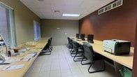 This single-floor space functions as a dedicated computer lab or business center, designed to accommodate multiple users in a structured, productive environment. The room features two long, light-wood workstation tables arranged in parallel rows, each equipped with desktop computers and ergonomic sled-base chairs, providing ample workspace for several individuals simultaneously. The layout is well-organized, promoting both focused individual work and group sessions. A printer or multifunction device is positioned at one end of the room, adding practical office functionality. The flooring consists of large, light-colored ceramic tiles that lend a clean and professional appearance throughout the space. Walls are painted in a two-tone scheme, with warm taupe on three sides and a deep, rich brown accent wall that adds visual depth and a modern contrast. The drop ceiling features recessed fluorescent panel lighting, ensuring consistent, even illumination across all workstations. A window fitted with horizontal blinds along one wall allows for natural light control while maintaining privacy. HVAC vents are visible in both the ceiling and upper wall, ensuring a comfortable climate year-round. Overall, the space conveys a functional, no-frills professional atmosphere suited for business or educational purposes.