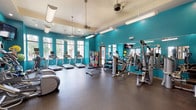 This fitness center is a thoughtfully designed workout facility that combines functionality with an energizing aesthetic. The space is defined by bold teal accent walls that create a vibrant, motivating atmosphere, complemented by a neutral rubber flooring that provides both durability and comfort underfoot. Elevated ceilings fitted with ceiling fans and a combination of pendant and recessed lighting ensure the space feels open and well-illuminated throughout.

Large windows along multiple walls flood the interior with natural light and offer views of the surrounding outdoor greenery, enhancing the overall workout experience. The cardio zone is well-stocked with multiple treadmills, elliptical trainers, and stationary bikes, each equipped with individual monitors for entertainment during workouts.

The strength training area features a multi-station cable machine, a dedicated free weight rack with a full range of dumbbells, and a flat workout bench. A wall-length mirror runs along one side, visually expanding the space while allowing users to monitor their form. Exercise balls and additional accessories are neatly organized on a storage rack nearby. A healthy vending machine offering snacks and drinks adds a convenient amenity, and a dedicated exit door provides easy outdoor access.