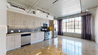 Located at 819 Santee St in the heart of downtown Los Angeles, this urban loft showcases a compelling blend of industrial character and modern refinement. The open-plan layout is anchored by gleaming polished concrete floors that reflect natural light throughout, while exposed ceiling infrastructure — including visible ductwork, conduit, and track lighting — reinforces the space's loft aesthetic.

The kitchen is thoughtfully designed with light wood cabinetry extending to the ceiling, a mosaic tile backsplash, a full suite of stainless steel appliances including a French door refrigerator, electric range, built-in microwave, and dishwasher. The kitchen opens directly to the main living area, fostering a seamless, connected feel.

Large factory-style windows flood the living space with abundant natural light and offer views of the surrounding downtown cityscape, framed by floor-length draping curtains. The main room features a frosted-glass sliding door that adds a sleek, contemporary partition element.

A hallway with a distinctive curved architectural column leads to the bathroom, which is well-appointed with a combined tub and shower enclosed by sliding glass doors, stone-textured tile surround, a wooden vanity, and large-format floor tiles. The entry features decorative marble-style tile flooring, adding an elegant touch to the industrial backdrop.