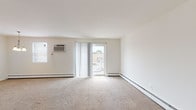 This single-floor unit offers a straightforward, functional layout across several well-defined spaces. The living area is generously proportioned, featuring neutral wall-to-wall carpeting, crisp white walls, and a sliding glass door fitted with vertical blinds that invites natural light and provides access to an outdoor area. A wall-mounted air conditioning unit and baseboard heating ensure year-round climate control throughout the carpeted rooms.

The kitchen is separated from the living area by a partial wall, creating a semi-open feel. It features white cabinetry with ample upper and lower storage, light countertops, a stainless steel sink, and white appliances including a gas range with oven and a full-size refrigerator. Hardwood-style flooring completes the kitchen space, offering a practical contrast to the carpeted areas.

The bedroom is a quiet, well-lit room with its own wall-mounted A/C unit, baseboard heating, and a window with blinds. The bathroom features a full-size soaking tub with tiled surround, and a clean, white aesthetic throughout. The overall ambiance of the unit is neutral and move-in ready, providing a clean canvas for personalization.