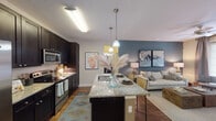 This single-floor residence offers a thoughtfully designed open-concept layout that seamlessly connects the kitchen, dining, and living areas. The kitchen is a standout feature, showcasing dark espresso cabinetry reaching toward the ceiling, granite countertops, stainless steel appliances including a built-in microwave and range, and a central island with a sink and pendant lighting overhead — ideal for both cooking and casual dining. A dedicated dining nook provides an intimate space for meals, accented by warm wood flooring, a round table with bench seating, and decorative wall art. The living room benefits from large windows that invite natural light, wood-style flooring, and a ceiling fan, creating a bright and comfortable gathering space. The bedrooms are carpeted for warmth and comfort, each featuring ceiling fans, windows with natural light, and ample room for furnishings. A full bathroom includes a soaking tub and shower combination with tile surround, dark cabinetry vanity, and wood-look flooring. Throughout the space, a neutral color palette with accent walls in muted tones provides a cohesive and contemporary aesthetic, complemented by consistent hardwood-style flooring in the main living areas.