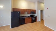 This single-floor studio with one bathroom offers an efficient, open layout that combines the living and kitchen areas into one cohesive space. The living area is characterized by light hardwood flooring and notably tall ceilings that lend a sense of openness uncommon in studio configurations. Large floor-to-ceiling windows along one wall allow generous natural light to fill the space, with what appears to be a sliding glass door providing direct outdoor access and views of surrounding greenery.

The kitchen runs along one wall and is well-equipped with warm-toned wood cabinetry both above and below the countertops. Black appliances, including a full-size refrigerator, electric range, built-in microwave, and dishwasher, are neatly integrated beneath dark countertops, providing a functional contrast to the neutral walls.

The bathroom continues the clean aesthetic with a white vanity featuring under-sink storage, light tile flooring, and a glass-enclosed tub and shower combination. The tall walls extend into the bathroom, reinforcing the vertical spaciousness found throughout. A closet is accessible from the main living area, adding practical storage to this thoughtfully designed studio.