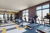This expansive fitness center is thoughtfully designed to accommodate a full range of workout routines within a stylish, modern environment. The space is organized into distinct functional zones, each tailored to a specific type of training. The cardio area lines the perimeter near floor-to-ceiling windows framed in sleek black metal, flooding the space with natural light and offering elevated outdoor views. Treadmills, ellipticals, stationary bikes, and assault bikes are positioned to take full advantage of this bright, airy setting.

The strength training zone features a robust free weights section with a tiered dumbbell rack, a weight bench, and a multi-functional squat and barbell rack. Large mirrors expand the sense of space and assist with form, while wall-mounted smart TVs add an entertainment dimension. A functional training area includes a cable machine, resistance bands, medicine balls, and a stability ball rack for versatile cross-training options.

Design details elevate the space beyond a standard gym. Decorative wallpaper with bold botanical and feather motifs, wainscoted walls in deep plum and soft grey, and a geometric patterned carpet in gold, charcoal, and white create a cohesive, upscale aesthetic. A welcoming entry area with a vibrant magenta storage console adds a striking accent upon arrival.