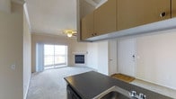 This single-floor residence offers an open and functional layout that seamlessly connects the kitchen and living areas. The kitchen is equipped with dark granite-style countertops, ample upper cabinetry in a warm wood finish, and a double-basin sink, providing a practical and well-appointed workspace. The space flows directly into the living area, which is finished with plush carpeting and features a built-in fireplace as a central focal point, adding warmth and character to the room. A ceiling fan overhead enhances comfort and air circulation throughout the main living space. Natural light fills the interior through a sliding glass door that opens to a private balcony, offering outdoor views and a connection to the outside. The entryway is defined by a clean white door with modern hardware, transitioning from hardwood-style flooring at the entry to the carpeted living area. The overall color palette is soft and neutral, with warm beige tones on the walls and cabinetry creating a cohesive and inviting ambiance. The thoughtful layout and mix of finishes make this a comfortable and well-designed single-level space.