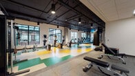 Located at 135 E 6th Ave in Eugene, Oregon, this well-appointed fitness center offers a comprehensive workout environment designed to accommodate a wide range of exercise routines. The main floor is expansive and open, featuring a striking geometric floor design in yellow, green, and white that adds a vibrant, energetic aesthetic to the space. Large industrial-style windows line the walls, flooding the facility with natural light while providing views of the surrounding exterior. Exposed black ductwork and pendant lighting run along the dark ceiling, giving the space a modern industrial character. The cardio zone is generously equipped with treadmills, ellipticals, rowing machines, and stationary bikes, all arranged to take full advantage of the natural light from the large windows. A functional training area includes TRX suspension training systems and cable machines, while full-length mirrors enhance spatial awareness and form feedback throughout. A separate free weight room offers rubber flooring, adjustable benches, and a complete dumbbell rack, with its own mirrored wall and access to additional rooms. The facility is thoughtfully designed to support cardio endurance, strength training, and functional fitness within a single, cohesive space.