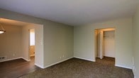 This single-floor residential space offers a functional and straightforward layout designed for comfortable everyday living. The interior is finished with wall-to-wall carpeting in a warm, neutral tone that adds a sense of coziness underfoot, while the surrounding walls are painted in a soft beige that promotes a calm and welcoming atmosphere throughout.

The main living area is open and generously proportioned, providing ample room for a variety of furniture arrangements. White baseboards frame the perimeter of the space, adding a clean, finished look that contrasts neatly with the neutral walls. A textured ceiling adds a subtle visual element overhead.

A built-in closet with bifold doors offers practical storage directly within the living space, making organization straightforward and convenient. An adjacent room, visible through an open doorway, features hard-surface flooring and benefits from natural light streaming in through a window, suggesting it may serve as an additional living or utility area.

The overall ambiance of the space is clean, understated, and move-in ready, with a neutral palette that provides a versatile backdrop for a range of interior styles. The simple, uncluttered design allows occupants to personalize the space with ease.