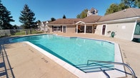 This community amenity space offers residents a well-rounded set of shared facilities designed for both relaxation and social engagement. At the heart of the outdoor area is a generously sized swimming pool with crystal-clear blue water, accessible via built-in stair rails and surrounded by a clean concrete pool deck. The pool area is framed by an ornamental metal fence and looks out toward a charming clubhouse building clad in light siding with a brown hip roof, multiple French-style glass doors, and a covered patio — all set against a backdrop of mature trees and open sky.

Adjacent to the pool, the clubhouse interior houses a multipurpose meeting and gathering room. This room features a drop tile ceiling with recessed fluorescent lighting, neutral textured wallcovering, and light tile flooring. It is furnished with a mix of rectangular conference-style tables and round tables, all accompanied by dark red cushioned chairs. The walls are adorned with a rotating selection of framed artwork, including landscape paintings and architectural photography, adding warmth to the functional space.

The building's interior corridors feature patterned carpeting in deep burgundy tones with ornate detailing, connecting the meeting spaces to additional rooms and exit points. The overall environment is clean, practical, and community-oriented.