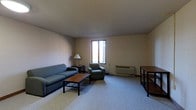 Located at 186 Mill St in Athens, Ohio, this single-floor space offers a straightforward and functional layout well-suited for everyday living. The main living area is finished with wall-to-wall carpeting in a neutral tone that complements the understated, light-colored walls and smooth ceiling above. A central window allows natural light to filter into the room, brightening the space and creating a welcoming atmosphere. The living area is furnished with a blue sofa and a matching armchair arranged around a wooden coffee table, providing a cozy seating arrangement. A floor lamp positioned near the seating area adds supplemental lighting. Along one wall, a wall-mounted HVAC unit ensures climate control throughout the space. A wooden shelving unit or side table is situated near the perimeter, offering additional storage or surface space. The overall design is clean and utilitarian, with a calm, neutral color palette throughout. The layout is open and uncluttered, making efficient use of the available floor space. This space is practical and comfortable, with essential amenities in place to support day-to-day living in the heart of Athens, Ohio.