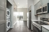 This single-floor unit offers a efficiently organized layout that makes excellent use of its compact footprint. The interior is finished in a neutral, contemporary palette of soft grays and warm whites throughout, creating a cohesive and clean aesthetic.

The kitchen serves as a standout feature, outfitted with a full suite of stainless steel appliances including an electric range, over-the-range microwave, refrigerator, and dishwasher. Gray shaker-style cabinetry extends from floor to ceiling, maximizing storage capacity. A crisp white subway tile backsplash complements the light quartz countertops, while dark hardwood-style flooring grounds the space with warmth and contrast.

The living area benefits from generous natural light, as evidenced by sunlight streaming across the neutral carpet, lending the space an open, airy quality. Elevated ceilings enhance the sense of volume throughout the unit. A closet with a rod and shelving system provides organized storage in the sleeping area.

The bathroom features gray cabinetry consistent with the kitchen's design, a single-sink vanity with a lighted mirror, and tile flooring. The unit also includes an in-unit washer and dryer, a balcony for outdoor access, and a well-connected, practical floor plan.