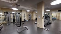 This fitness center offers a comprehensive workout environment designed to accommodate a range of exercise needs. The space features durable rubber flooring throughout, providing both safety and comfort during workouts. Mirrored walls line multiple sides of the room, enhancing the sense of openness while allowing users to monitor their form during exercise.

The facility is outfitted with a solid selection of strength training equipment, including cable machines, a multi-functional weight station, and free-standing resistance apparatus. Cardio equipment, such as an elliptical trainer and a treadmill, is positioned along the perimeter, offering users a well-rounded fitness experience. A BOSU balance trainer and additional functional fitness tools are also available for varied training routines.

Overhead pendant lighting illuminates the space evenly, while exposed ductwork along the ceiling contributes to an industrial aesthetic. Structural columns are integrated throughout the layout, defining zones within the room. The neutral, cream-toned walls create a clean, focused atmosphere suited for exercise. The overall design prioritizes functionality, making efficient use of the available floor space.