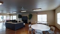 This single-floor multipurpose common room is designed to accommodate both relaxation and group gatherings. The space is divided into two functional zones that flow seamlessly into one another. On one side, a comfortable lounge area features a dark sofa and loveseat arranged around a large flat-screen television mounted on a media stand, creating an ideal setting for leisure and entertainment. On the other side, round white tables paired with folding chairs provide ample seating for dining or group activities. The room is finished with warm terra cotta tile flooring throughout, contributing to a grounded, welcoming atmosphere. Overhead, flush-mount fluorescent light fixtures and a ceiling fan ensure the space is well-lit and ventilated. Large windows along the walls allow natural light to filter in, brightening the interior. Tall potted indoor trees are positioned throughout the room, adding a touch of greenery and life to the space. The walls are finished in a neutral tone, and the overall layout is open and functional, making it well-suited for a variety of communal uses.