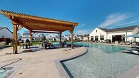 Located at 9944 Highway 6 in Missouri City, Texas, this single-level outdoor community amenity space is thoughtfully designed to provide residents with a resort-style experience. At the heart of the space is a generously sized swimming pool with crystal-clear water, highlighted by a standout in-pool island pergola constructed of warm wood timber. The pergola shelters a seating and dining area, creating a shaded retreat accessible directly from within the pool — ideal for relaxed gatherings. Surrounding the pool, an expansive concrete sun deck is lined with cushioned lounge chairs and striped umbrellas, offering ample space for sunbathing and leisure. A separate spa provides an additional relaxation option adjacent to the main pool. The amenity area also features two distinct lawn zones covered in lush artificial turf. One lawn area is set up for lawn games, including cornhole boards and casual seating beneath string lights, while the other offers a ping pong table, a hanging pod chair, and open play space. String lights strung between poles create a warm, festive ambiance throughout the grounds. The white-sided clubhouse building frames one side of the space, with the surrounding residential neighborhood visible beyond the black iron perimeter fencing, giving the area a welcoming, community-oriented character.