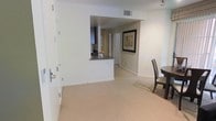 This single-floor residence offers a clean, functional layout designed to maximize comfort and flow. The living and dining area is finished with plush wall-to-wall carpeting in a warm neutral tone, creating a welcoming atmosphere that complements the bright white walls throughout. A pass-through opening connects the kitchen to the main living space, providing an open, communicative feel between rooms. The kitchen features dark cabinetry and granite-style countertops visible through the pass-through, adding a touch of contrast against the light surroundings. A hallway extends from the main living area, leading to additional rooms and offering a clear sense of organized spatial separation. Recessed and track lighting provides overhead illumination, while HVAC venting integrated into the ceiling ensures climate comfort year-round. The dining area is positioned conveniently near the kitchen pass-through, accommodating a dining set with ease. Framed wall art and white baseboards throughout contribute to a polished, move-in-ready aesthetic. The overall ambiance is bright, neutral, and understated — well-suited for a variety of interior styles and furnishing preferences.