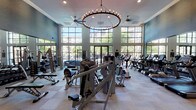 Located at 2121 Delgany Street in Denver's vibrant Lower Downtown neighborhood, this fitness center is a standout amenity defined by its dramatic double-height ceilings and floor-to-ceiling windows that flood the space with natural light and offer views of the surrounding greenery. The open, airy layout is anchored by a striking circular candle-style chandelier and complemented by large ceiling fans that keep the environment comfortable during workouts. The design balances industrial and contemporary aesthetics, featuring textured dark accent walls, reclaimed wood paneling that extends toward the ceiling, and large wall-mounted mirrors that visually expand the space. The strength training zone is outfitted with a comprehensive selection of Hoist cable machines, free weights, adjustable benches, and multi-functional racks. The cardio area lines the perimeter and includes Cybex treadmills, rowing machines, ellipticals, and upright bikes, each equipped with personal monitors. Wall-mounted televisions provide entertainment throughout. Additional amenities include storage cubbies, exercise mat storage, and convenient pass-through access to adjacent areas. The flooring transitions between carpet tiles and wood-look surfaces, delineating functional zones within the space. Overall, the fitness center delivers a polished, professional environment suited for a full range of training activities.
