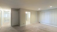This single-floor residential space offers a clean, functional layout with a straightforward and practical design. The main living area is finished with wall-to-wall carpeting in a warm neutral tone, providing a comfortable and quiet underfoot feel. The walls throughout are painted in a soft, light beige, creating a calm and understated ambiance that allows for easy personalization.

A wide, open doorway connects the living area to an adjacent room, which features hard-surface flooring and a window that allows natural light to filter through, brightening both spaces. A set of vertical blind-covered sliding glass doors lines one wall of the living area, offering potential access to an outdoor area while providing light control and privacy.

Additional features include a closet with a sliding door, a recessed ceiling light, and wall outlets positioned conveniently throughout. The white-painted trim and baseboards add a crisp contrast to the neutral walls, lending a tidy, well-maintained appearance. The overall layout is open and uncluttered, making it an adaptable space suited for a variety of living arrangements. The home's single-floor configuration ensures easy accessibility and a cohesive flow throughout.