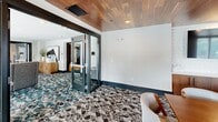 Located at 5580 Farrington Rd in Durham, NC, this sophisticated single-floor amenity suite offers a thoughtfully designed layout across three distinct areas, each with its own character and purpose.

The main lounge serves as the heart of the space, anchored by a striking geometric LED chandelier and floor-to-ceiling glass railings that flood the room with natural light and offer expansive views of the surrounding area. Plush high-back lounge chairs are arranged around a sleek black coffee table atop a bold teal and ivory patterned carpet, creating an inviting atmosphere ideal for casual gatherings or collaborative work.

Glass-paneled doors connect the lounge to a private conference room, finished with a warm wood-plank ceiling that adds a refined, organic touch. A large oval conference table surrounded by modern upholstered chairs makes this room well-suited for meetings and presentations. Framed geometric artwork adorns the walls, complementing the contemporary aesthetic, while generous windows maintain a connection to the outdoors.

A third, more intimate room features the same rich wood ceiling, wall-mounted display screen, built-in wood cabinetry, and a compact seating area with stylish lounge chairs in soft grey and purple tones. Patterned carpeting flows throughout, tying all three spaces together. Black-framed glass doors and crisp white walls complete the polished, modern design of the suite.