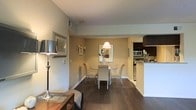 This single-floor space showcases an open-concept layout that seamlessly connects the living, dining, and kitchen areas into one cohesive environment. Dark hardwood flooring runs throughout, providing a rich contrast against the warm beige and soft grey wall tones that define the interior palette.

The dining area serves as a central focal point, furnished with a round glass-top table and upholstered chairs positioned beneath a classic pendant light, creating an intimate yet functional setting. A framed mirror and wall art add decorative character to the surrounding walls.

Adjacent to the dining space, the kitchen is equipped with stainless steel appliances, dark cabinetry, and a white countertop peninsula that provides both workspace and a natural visual boundary between the cooking and living zones. An overhead range hood and built-in microwave reflect a practical, well-appointed layout.

The living area, partially visible in the foreground, features a side table with a contemporary lamp and a decorative mirror, contributing to the space's refined, understated ambiance. Recessed lighting and a textured ceiling add subtle architectural detail throughout.