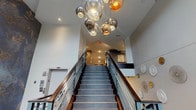 This well-appointed building lobby makes a strong first impression with its dramatic entrance staircase, framed by sleek metal and wood railings and crowned by a sculptural cluster of globe pendant lights in smoked and metallic finishes. The soaring ceiling height of the entryway amplifies the sense of arrival and adds a refined, architectural character to the space.

The main lobby area transitions seamlessly into a communal lounge zone, where rich, warm-toned hardwood flooring contrasts elegantly with gray carpet accents. A high-top communal table paired with upholstered bar-height chairs sits beneath a pair of industrial-style pendant lights, offering residents a casual gathering or working space. Abstract artwork adorns the walls, adding a creative, contemporary touch to the overall aesthetic.

A dedicated mail center is a standout functional feature, lined wall-to-wall with an extensive bank of individual resident mailboxes, finished in brushed aluminum and set against dark wood trim. The area is thoughtfully appointed with a small counter for sorting mail and personal convenience items.

Connecting corridors are finished in neutral tones with gray carpeting and clean white walls, maintaining a calm and consistent design language throughout. The space balances modern style with practical amenities, creating a welcoming and efficient environment for building residents.