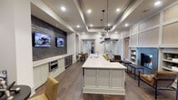This single-story space showcases a thoughtfully designed open-concept interior that seamlessly blends functional and social areas. At its center sits a generous white kitchen island with a clean quartz surface, overhead pendant lighting in a clustered lantern style, and an integrated sink — serving as the focal point of the space.

The kitchen zone features white cabinetry, stainless steel appliances including a wall oven and beverage coolers, and a dramatic textured dark stone accent wall mounted with a flat-screen television. This media wall adds a bold contrast to the otherwise light and neutral palette throughout the space.

Tray ceilings with recessed lighting lend architectural depth and a refined character to the room, while wide-plank hardwood flooring runs continuously, anchoring the design with warmth. A built-in shelving unit with a fireplace surround occupies one wall, adding a cozy residential touch.

Seating areas with upholstered chairs are positioned throughout, creating comfortable gathering zones. French doors at the rear allow for visual connection to adjoining spaces, contributing to the open and airy ambiance that defines this well-appointed interior.