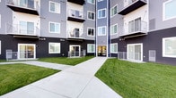 This modern residential building showcases a striking exterior defined by a contemporary color palette of deep navy blue, crisp white, and dark charcoal siding. The multi-story structure is designed around a central courtyard, which serves as a shared outdoor amenity for ground-floor residents. The courtyard features neatly manicured green lawns divided by clean concrete walkways, creating an organized and inviting outdoor environment.

Ground-floor units open directly onto the courtyard via sliding glass doors, each with a private patio enclosed by sleek metal railings, offering a seamless indoor-outdoor connection. Large windows throughout the building allow for generous natural light across all levels. Upper-floor units are equipped with private balconies framed by metal railings, providing elevated outdoor space with views of the surrounding area.

The building's facade combines horizontal lap siding with bold accent panels, giving it a distinctly modern aesthetic. The overall design balances privacy with a sense of community, as the courtyard layout encourages an open, connected atmosphere. The well-maintained landscaping and clean architectural lines contribute to a polished, contemporary ambiance throughout the property.