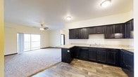 This single-floor space is designed with an open-concept layout that seamlessly connects the kitchen and living area, creating a spacious and cohesive environment. The kitchen is a standout feature, showcasing rich, dark espresso-toned cabinetry that extends from base to upper wall units, providing generous storage throughout. Light-toned countertops offer a striking contrast against the dark cabinetry, while a double sink is conveniently positioned beneath recessed lighting. A built-in dishwasher is integrated into the lower cabinetry for a clean, streamlined look. The kitchen transitions naturally into the living area by way of a peninsula that offers additional counter space and serves as a natural divider between the two zones. Durable hardwood-style flooring runs through the kitchen area, while plush wall-to-wall carpeting defines the living space, adding warmth and comfort underfoot. The living area is outfitted with a ceiling fan, enhancing air circulation throughout. A sliding glass door at the far end of the living area allows natural light to pour in and provides access to an outdoor balcony, connecting the interior to the outside. The overall ambiance is clean, modern, and move-in ready.