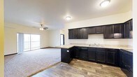 This single-floor space is designed with an open-concept layout that seamlessly connects the kitchen and living area, creating a spacious and cohesive environment. The kitchen is a standout feature, showcasing rich, dark espresso-toned cabinetry that extends from base to upper wall units, providing generous storage throughout. Light-toned countertops offer a striking contrast against the dark cabinetry, while a double sink is conveniently positioned beneath recessed lighting. A built-in dishwasher is integrated into the lower cabinetry for a clean, streamlined look. The kitchen transitions naturally into the living area by way of a peninsula that offers additional counter space and serves as a natural divider between the two zones. Durable hardwood-style flooring runs through the kitchen area, while plush wall-to-wall carpeting defines the living space, adding warmth and comfort underfoot. The living area is outfitted with a ceiling fan, enhancing air circulation throughout. A sliding glass door at the far end of the living area allows natural light to pour in and provides access to an outdoor balcony, connecting the interior to the outside. The overall ambiance is clean, modern, and move-in ready.