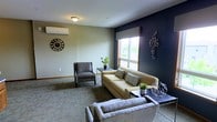 This single-floor space offers a thoughtfully designed interior that combines modern aesthetics with a comfortable, inviting atmosphere. The lounge area serves as the centerpiece of the space, featuring a cohesive mix of furnishings including a plush tan sofa accented with patterned throw pillows, a coordinating armchair, and additional seating arranged for easy conversation and relaxation. A bold navy blue accent wall provides a striking visual contrast against the neutral beige walls, adding depth and a contemporary edge to the room's overall design. Decorative wall art and greenery throughout contribute to a polished yet approachable feel. Natural light filters in through a large window with a roman shade, brightening the carpeted interior and offering a view to the outdoors. A wall-mounted climate control unit ensures year-round comfort. Subtle finishing details, including baseboards and carefully selected décor such as a large wall clock and geometric accent pieces, reflect a consistent attention to design. The layout is efficient and well-organized, making the space functional for both social gatherings and quiet use.