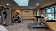 This fitness center offers a thoughtfully designed workout space finished with warm beige walls accented by white chair rail molding and crown molding details. A prominent overhead skylight floods the room with natural light, complementing the recessed lighting installed throughout the ceiling. The flooring consists of durable, patterned carpet tiles suited for high-traffic athletic use.

The space is equipped with multiple Matrix-brand treadmills and elliptical trainers, providing ample cardio options. A multi-function cable resistance station is positioned in one corner, supporting strength and functional training exercises. A wall-mounted flat-screen television is positioned for easy viewing during workouts. A large floor-to-ceiling mirror spans one wall, visually expanding the space while allowing users to monitor their form.

Climate comfort is ensured by a wall-mounted ductless mini-split air conditioning unit, along with a visible thermostat for temperature control. A fire extinguisher and an AED defibrillator are on hand for safety. Natural light also enters through traditional multi-pane windows, offering views of the surrounding exterior. The single-floor layout and efficient use of space make this fitness center a practical and well-appointed facility.