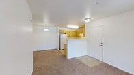 This single-floor, 3-bedroom, 2-bathroom residence offers a well-designed layout that maximizes comfort and functionality. The open-concept living and dining area features neutral wall-to-wall carpeting that creates a warm, inviting atmosphere, while flush-mount and pendant lighting fixtures provide consistent illumination throughout the space. A peninsula-style kitchen is positioned at the heart of the home, featuring warm-toned wood cabinetry, a white refrigerator, and a raised breakfast bar that serves as a natural divider between the kitchen and living areas. The kitchen's tile flooring transitions smoothly from the carpeted living space, adding a practical touch to the cooking area. The layout promotes an easy flow between rooms, making the space ideal for both everyday living and entertaining. The white interior walls and ceilings contribute to a clean, bright aesthetic throughout, while the front entry door with hardware provides a welcoming point of arrival. With three bedrooms, the home offers generous flexibility for families, guests, or a dedicated home office, all contained within a compact and efficiently organized single-level floor plan.