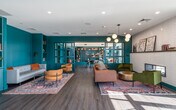 This thoughtfully designed single-floor community space blends a library, lounge, and social gathering area into one cohesive and visually striking environment. The space is defined by its bold teal color palette, which is carried throughout built-in bookshelves, accent walls, and architectural millwork, creating a warm yet energetic atmosphere.

The library area features floor-to-ceiling teal built-in shelving stocked with books and decorative objects, complemented by round wood tables and modern chairs suited for reading or collaborative work. A dedicated nook with a wall-mounted display screen and seating offers a more intimate workspace within the larger room.

The lounge area provides a comfortable retreat anchored by a leather sofa, green accent chairs, and a patterned area rug. A sleek linear fireplace set into a white textured tile surround serves as a focal point, while a contemporary multi-globe pendant chandelier adds a stylish touch overhead. Curated botanical artwork adorns the walls, reinforcing the space's refined yet approachable character.

Large floor-to-ceiling windows and glass doors flood the interior with abundant natural light and offer open views of an outdoor terrace equipped with additional seating. The wood-look flooring and coffered ceiling with integrated ambient lighting tie the entire space together, resulting in a polished, inviting environment well-suited for both focused work and relaxed socializing.