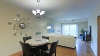 This single-floor space offers an open-concept layout that seamlessly connects the dining and living areas, creating a cohesive and functional environment. The dining area is anchored by a round marble-top table surrounded by dark leather chairs, topped with an elegant multi-arm chandelier that serves as a striking focal point. A decorative starburst mirror adorns the soft blue-green wall, adding a contemporary artistic touch to the space.

The living area flows naturally from the dining space and features a plush white sectional sofa, complemented by a tall arched floor lamp and a coordinating side table lamp. A framed blue artwork on the wall adds a pop of color and visual interest. The space is bathed in natural light from large sliding glass doors that open to an outdoor balcony, offering views of lush surrounding greenery.

Warm hardwood flooring runs throughout both areas, tying the space together with a sense of continuity and warmth. The light-colored walls and abundant natural light contribute to an airy, open ambiance. Ceiling vents indicate a centralized climate control system, ensuring year-round comfort. Overall, the space strikes a balance between modern design and approachable comfort.