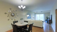 This single-floor space offers an open-concept layout that seamlessly connects the dining and living areas, creating a cohesive and functional environment. The dining area is anchored by a round marble-top table surrounded by dark leather chairs, topped with an elegant multi-arm chandelier that serves as a striking focal point. A decorative starburst mirror adorns the soft blue-green wall, adding a contemporary artistic touch to the space.

The living area flows naturally from the dining space and features a plush white sectional sofa, complemented by a tall arched floor lamp and a coordinating side table lamp. A framed blue artwork on the wall adds a pop of color and visual interest. The space is bathed in natural light from large sliding glass doors that open to an outdoor balcony, offering views of lush surrounding greenery.

Warm hardwood flooring runs throughout both areas, tying the space together with a sense of continuity and warmth. The light-colored walls and abundant natural light contribute to an airy, open ambiance. Ceiling vents indicate a centralized climate control system, ensuring year-round comfort. Overall, the space strikes a balance between modern design and approachable comfort.
