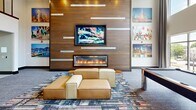Located on Campus Circle in Urbana, Illinois, this dynamic two-floor amenity facility offers a wide range of recreational and social spaces designed for an active, community-oriented lifestyle. The ground floor welcomes visitors with a vibrant lounge area characterized by bold pop-art wall installations, patterned wallcoverings, and eclectic game tables including poker and mahjong setups, all accented by a built-in beverage cooler. A fully appointed communal kitchen and lounge area features a long central island with bar seating, rich wood cabinetry, pendant lighting, and integrated flat-screen televisions, alongside cozy booth-style seating areas framed by decorative tile wall accents. Exposed brick walls and wood-look flooring add warmth throughout. The upper floor transitions to a more active atmosphere, housing a generously proportioned fitness center equipped with cardio machines and treadmills, floor-to-ceiling windows that flood the space with natural light and offer open views of the surrounding streetscape. Large wall mirrors enhance the sense of space. A game lounge with ping-pong tables, exposed brick walls, mounted televisions, and vibrant artwork completes the upper level. A dedicated cycle studio and additional lounge areas with workstations round out this well-appointed amenity space.