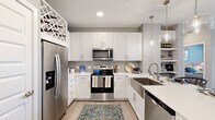 This thoughtfully designed single-floor residence offers a cohesive blend of modern aesthetics and functional living. The kitchen serves as a centerpiece, featuring crisp white shaker cabinetry, a grey tile backsplash, and a full suite of stainless steel appliances including a french door refrigerator with ice dispenser, an electric range with over-the-range microwave, and a dishwasher. Light quartz countertops extend to a spacious island with seating for multiple bar stools, illuminated by elegant glass pendant lights. Open shelving along one wall provides additional display and storage space, while a decorative lattice wine rack adds a charming architectural touch above the cabinetry. Light wood-tone flooring flows throughout the main living areas, lending warmth and continuity to the space. The living room is bright and airy, featuring a ceiling fan, large windows dressed with linen drapes, and a relaxed, neutral color palette that creates a calm, welcoming atmosphere. A hallway connects the main living spaces to the bedroom, which features plush carpet, generous natural light through a wide window, soft blue-grey walls, and a serene ambiance well-suited for rest. The overall design emphasizes clean lines, neutral tones, and practical elegance throughout.