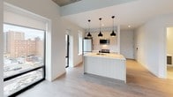 This single-floor unit showcases a thoughtfully designed modern interior with clean lines and a neutral palette throughout. The open-concept living and kitchen area serves as the heart of the space, featuring light wood-toned flooring that flows seamlessly from room to room. The kitchen is well-appointed with light wood cabinetry, a large central island with a white countertop, stainless steel appliances including a refrigerator and microwave, and a trio of sleek pendant lights overhead. Multiple windows flood the kitchen and living area with natural light while offering elevated views of the surrounding urban cityscape.

The bedroom features exposed concrete ceiling details, adding an industrial character to the otherwise minimalist design. A full-height sliding glass door provides additional natural light and outdoor views. The bathroom is finished with a frameless glass walk-in shower, light subway-style wall tiling, mosaic tile flooring, and polished chrome fixtures, creating a clean, spa-like atmosphere. A dedicated utility area houses a stacked washer and dryer unit alongside a water heater, keeping all mechanical elements neatly contained. Overall, the space presents a cohesive contemporary aesthetic with practical, well-integrated features throughout.