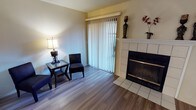 This single-floor space offers a welcoming and functional layout designed for comfortable everyday living. The living area serves as the centerpiece of the home, showcasing warm hardwood-style flooring that runs throughout and lends a clean, cohesive aesthetic to the space. A tile-surround fireplace with a classic white mantel provides a focal point in the room, adding both architectural character and a cozy ambiance. Decorative accents atop the mantel enhance the inviting atmosphere. Natural light fills the living area through a large sliding glass door fitted with vertical blinds, offering both privacy and easy access to the outdoors. The seating arrangement is intimate and practical, with accent chairs and a side table creating a relaxed conversation nook. The warm, neutral wall tones complement the flooring and trim throughout, contributing to a bright and airy feel. The overall design balances simplicity with comfort, making the space feel approachable and well-appointed. The single-floor layout ensures easy navigation and accessibility throughout the home.