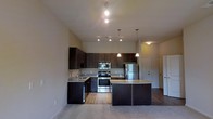 This single-floor space is designed with a modern aesthetic and an open-concept layout that promotes a sense of spaciousness and flow. The kitchen serves as the centerpiece, showcasing rich dark wood cabinetry that extends nearly to the ceiling, maximizing storage while adding a bold contrast to the neutral-toned walls. Stainless steel appliances, including a refrigerator, range, and built-in microwave, provide a sleek, contemporary finish. A curved kitchen island with light granite countertops offers additional workspace and serves as a natural gathering point, complemented by stylish pendant lighting that hangs overhead. Track lighting along the ceiling further illuminates the space, highlighting the tall ceiling height that contributes to an airy, open atmosphere. The flooring transitions from warm hardwood in the kitchen area to plush carpet in the adjacent living space, adding texture and comfort underfoot. The open layout allows the kitchen to flow seamlessly into the living area, making the overall floor plan feel connected and well-suited for both everyday living and entertaining. A private entry door is visible, adding a sense of arrival and separation to the space.