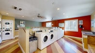 Located in Phoenix, Arizona, this shared laundry facility offers a practical and well-organized space designed to serve a residential community. The room is immediately distinguished by its eye-catching striped vinyl flooring in bold shades of terracotta, gold, and cream, lending the space a lively, energetic character. The walls are painted in a warm combination of burnt orange, sage green, and tan, creating a welcoming atmosphere that stands apart from a typical utilitarian laundry room.

The facility is outfitted with a row of front-loading washing machines and a bank of stacked dryers, providing ample laundry capacity for residents. A utility sink offers additional convenience for hand-washing or pre-treating garments. Granite-topped folding counters are positioned throughout the room, giving users dedicated surfaces for sorting and folding laundry. A card-operated payment station is mounted on the wall for easy access.

Natural light enters through multiple sliding windows that frame views of the surrounding residential buildings, keeping the space bright and airy. Recessed lighting and ceiling fans with integrated lights provide additional illumination and air circulation. Instructional postings and best-practice guides are displayed on the walls to assist users. The textured white ceiling and clean, well-maintained finishes throughout contribute to a tidy, functional environment.