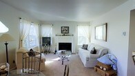 This single-floor space exudes a warm, inviting ambiance centered around a classic white-mantled fireplace that serves as the focal point of the living area. The room is finished in soft, neutral tones — pale walls and light carpeting — that create a calm and airy atmosphere throughout. Large windows dressed with sheer white curtains line the walls, allowing generous amounts of natural light to fill the space and enhance its open, comfortable feel. The living area is thoughtfully arranged with plush white slipcovered sofas accented by decorative pillows, surrounding a wrought-iron and glass coffee table that adds a touch of elegance without overwhelming the space. A wicker accent chair and decorative baskets introduce subtle coastal and natural textures, while a framed mirror and artwork complete the refined yet relaxed aesthetic. A television and media stand are positioned conveniently near the fireplace wall, blending function with style. Decorative pieces along the mantel add personality and charm to the space. The overall design balances casual comfort with tasteful detailing, making this a well-appointed living environment suited for both relaxing and entertaining.