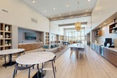 Located in Goodyear, Arizona, this thoughtfully designed single-floor community space offers a generous, open-concept layout that seamlessly integrates a fully equipped kitchen and an expansive living and lounge area. Soaring double-height ceilings throughout amplify the sense of space, while large floor-to-ceiling windows flood the interior with abundant natural light and frame views of the outdoor pool area and surrounding palm trees.

The kitchen features warm wood cabinetry, a striking marble-veined island with waterfall countertops, a decorative tile backsplash, open shelving, and modern appliances including a beverage refrigerator. Woven pendant lights add a natural, textural element above the dining counter and seating area.

The living room is anchored by a recessed accent wall with a mounted flat-screen TV and custom wood cabinetry below, complemented by plush neutral sofas accented with teal and blue pillows. A pool table serves as a central recreational feature. Multiple seating arrangements, including lounge chairs and round marble-top tables, offer versatile gathering options. Built-in display shelving and curated décor add visual interest throughout. Light wood-look flooring flows consistently across the entire space, contributing to its cohesive, contemporary aesthetic.