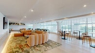 This thoughtfully designed single-floor space combines functionality with contemporary style across a series of interconnected zones. The open-plan layout is anchored by a spacious lounge area furnished with a distinctive sculptural sectional sofa in warm blush tones, set atop a bold botanical-patterned area rug. A built-in banquette with soft accent lighting, open shelving with greenery, and a wall-mounted display screen enhance the relaxed ambiance of this seating zone.

Adjacent to the lounge, a sleek full kitchen features an expansive stone-topped island with bar seating, white flat-panel cabinetry, stainless steel ovens, and a curated gallery wall of framed artwork. Pendant lighting with globe fixtures adds a warm, contemporary touch overhead. Light hardwood flooring runs throughout, contributing to the bright, cohesive aesthetic.

A dining area with dark tables and mixed seating connects the kitchen to the lounge, while a separate dining nook positioned alongside the large gridded windows offers views to the outdoors. A long corridor lined with a patterned runner leads to additional workspaces and private offices visible through glass partitions.

Floor-to-ceiling arched windows along the perimeter bathe the entire space in abundant natural light, creating an airy, welcoming environment well-suited for collaboration, gathering, or focused work.
