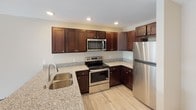 This single-floor unit offers a clean, modern aesthetic with consistent light hardwood-style flooring and neutral tones carried throughout every room. The kitchen is a standout feature, showcasing rich dark-stained wood cabinetry, granite countertops, and a full suite of stainless steel appliances including a refrigerator, range, and microwave. A peninsula with an undermount sink provides additional prep space and a natural transition into the adjacent living area, which benefits from recessed lighting overhead. The living space is bright and open, featuring a ceiling fan with light fixture and a window that draws in natural light while offering views of the surrounding greenery outside. Dark wood trim and baseboards provide a grounding contrast against the light walls and flooring, adding a sense of warmth and definition to each room. The bathroom continues the cohesive design language with matching dark cabinetry, granite countertops, an undermount sink, and a full tub-shower combination. The layout, as seen from the dollhouse view, is efficiently organized with well-defined spaces that flow naturally from one to the next, making the unit feel functional and comfortable throughout.