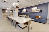 This contemporary office space, situated at 21 Barker Ave in White Plains, NY, showcases a thoughtfully designed 1,370 square foot single-floor layout. The interior is characterized by its open-concept design, creating a sense of spaciousness and fluidity throughout. Large windows flood the area with natural light, offering picturesque views of the surrounding landscape.

The space features a harmonious blend of work and relaxation areas. A striking reception area welcomes visitors, leading to various functional zones. The main work area includes an expansive countertop with bar-style seating, ideal for collaborative work or casual meetings. Adjacent to this is a comfortable lounge area with plush seating, perfect for breaks or informal discussions.

The color palette is predominantly neutral, with crisp white walls and ceilings contrasted by occasional accent walls in deep blue. This creates a calming yet professional atmosphere. The flooring consists of large, light-colored tiles that contribute to the overall brightness of the space.

Lighting is a key feature, with a combination of recessed ceiling lights, pendant fixtures, and LED strip lighting along the walls, providing both ambient and task lighting. The kitchen area is equipped with modern appliances and ample counter space, catering to employee needs.

Throughout the office, carefully placed plants add touches of greenery, enhancing the overall ambiance and air quality. The thoughtful integration of aesthetics and functionality makes this space an ideal environment for modern, dynamic businesses.