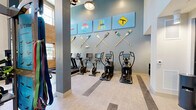 This fitness center offers a generously sized, single-floor exercise space designed to accommodate a comprehensive workout experience. The room's most striking architectural feature is its soaring double-height ceiling, which creates an open, airy atmosphere and is complemented by exposed brick accent walls that add an industrial character. Large windows positioned at both standard and elevated heights flood the space with abundant natural light, enhancing the energetic ambiance.

The flooring is finished in a light, durable material with contrasting stripe inlays that define different workout zones throughout the room. The cardio section is lined with treadmills, elliptical trainers, and stationary cycling bikes — all from the True Fitness brand — providing a robust selection for aerobic training. A dedicated strength area features a multi-station cable machine, a power rack, adjustable benches, a full dumbbell rack, and an assortment of medicine balls and stability equipment.

Full-length mirrors along one wall visually expand the space and assist with form during exercise. Distinctive pendant light fixtures with an industrial aesthetic add stylish overhead illumination. Decorative oar and paddle wall art, along with colorful artwork panels, contribute a vibrant, sporty personality to the interior. Built-in shelving cubbies near the entrance provide convenient storage, and a glass-paneled door offers a refined transition point within the facility.