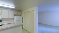 This single-floor space presents a straightforward and practical layout connecting the kitchen and living areas. The kitchen is equipped with light wood cabinetry mounted above the counter space, a white refrigerator, and a light-toned countertop that extends to form a partial divider between the kitchen and the adjacent area. The flooring in the kitchen zone is a light-colored vinyl or tile, providing a clean and easy-to-maintain surface. A bi-fold closet with matching light wood panel doors and gold-toned hardware is positioned along a dividing wall, offering convenient storage. The living area beyond features wall-to-wall carpet in a neutral tone, contributing to a softer, more comfortable atmosphere. The walls throughout are finished in a consistent off-white, keeping the overall look cohesive and understated. Overhead lighting is present in the kitchen area, providing functional illumination. The open connection between the kitchen and living space allows for a sense of continuity, making the overall layout feel accessible and easy to navigate.