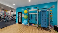This single-level fitness center is thoughtfully divided into distinct workout zones, each designed to support a comprehensive exercise experience. The main strength and cardio area features plush carpet flooring, recessed lighting, and a striking full-wall mural with bold, multicolored abstract artwork that energizes the space. This zone houses a selection of strength machines, ellipticals, treadmills, and a wall-mounted television, complemented by a ceiling fan for added comfort.

An adjacent functional training studio features light hardwood-style flooring and is anchored by a vibrant teal accent wall. Mounted to this wall is a Smart modular fitness rig equipped with TRX suspension straps, battle ropes, and resistance bands. Wall-mounted stability balls in varying sizes and a full rack of kettlebells provide additional versatility for functional and cross-training workouts.

A separate studio space, finished with the same hardwood-style flooring, is bright and open with multiple windows offering views of lush outdoor greenery. This room includes a Peloton cycle, a rowing machine, a large floor-to-ceiling mirror, a wall-mounted television, and a ceiling fan, making it ideal for cycling classes or individual cardio sessions. Direct access to an outdoor area further enhances the overall fitness experience.